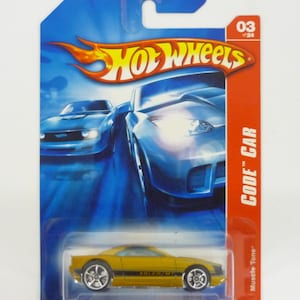 Peut inclure: Une voiture miniature Hot Wheels Code Car jaune en métal moulé sous pression avec le texte "ADLER/MT" sur le côté. La voiture est sur une carte bleue et rouge avec le logo Hot Wheels et le texte "Muscle Tone" et "Code Car".