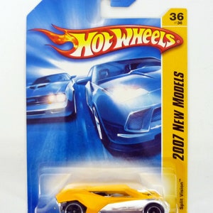 Puede incluir: Un coche de juguete Hot Wheels amarillo con una franja plateada, etiquetado como "Split Vision", en una tarjeta azul y amarilla con el texto "2007 New Models" y "36 de 36".