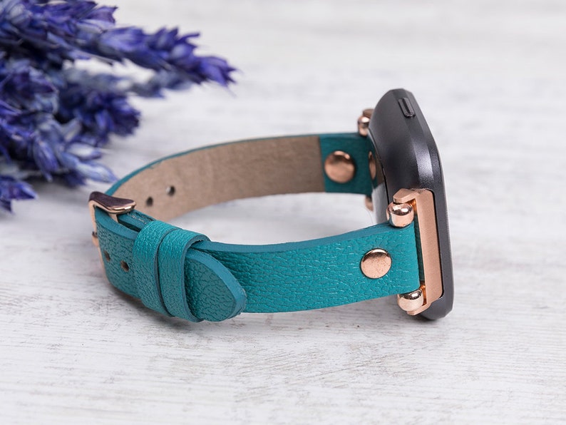May include: Teal leather watch strap with rose gold hardware and a black watch face.