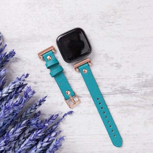 May include: A teal blue leather watch strap with rose gold hardware and a black square watch face.