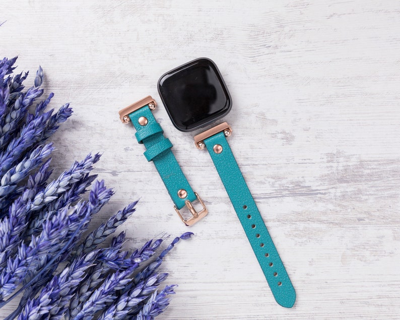 May include: A teal blue leather watch strap with rose gold hardware and a black square watch face.