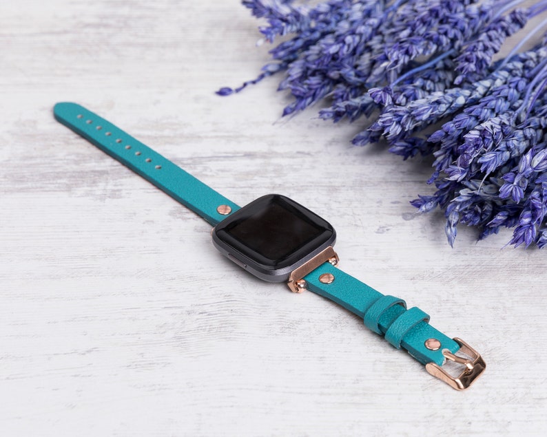 May include: A teal blue leather watch strap with rose gold hardware. The strap is designed for a smartwatch and has a minimalist style.