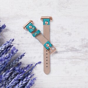 May include: Two teal leather watch straps with rose gold hardware. The straps are lying on a white wooden surface.