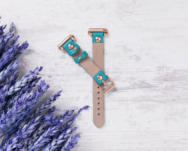 May include: Two teal leather watch straps with rose gold hardware. The straps are lying on a white wooden surface.