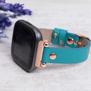 May include: A teal leather watch strap with rose gold accents and a black smartwatch face.