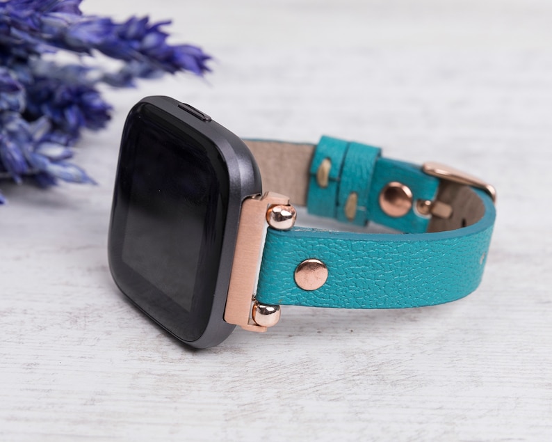 May include: A teal leather watch strap with rose gold accents and a black smartwatch face.