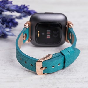May include: Teal leather watch strap with rose gold hardware and a black smartwatch face. The strap has a textured finish and is designed for a Fitbit Versa.