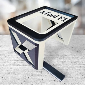 May include: A white and black 3D printed tool holder with the text "XTool F1" printed on the top. The holder has a rectangular base and a raised platform with a square opening.