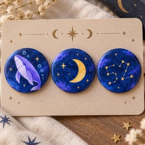 May include: Three circular pin-back buttons attached to a rectangular beige display card. The buttons feature dark blue celestial designs: a purple whale with bubbles, a yellow crescent moon, and a yellow star constellation forming the number 52.