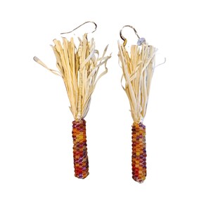May include: A pair of handmade earrings featuring a corn-on-the-cob design. The earrings have a beaded, cylindrical base in shades of red, orange, and yellow, topped with a cluster of beige raffia. The ear wires are silver-colored.