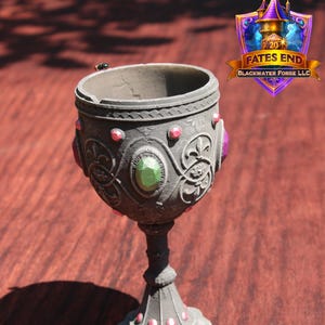 Könnte beinhalten: Ein dekorativer Kelch mit dunkelgrauer Oberfläche, verziert mit roten und grünen Edelsteinen. Der Kelch hat eine verzierte Detaillierung und einen ausgestellten Sockel, ideal für Fantasy-Dekorationen.
