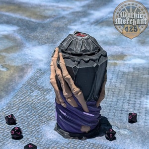 May include: A black and purple skeletal hand-shaped container with a red star on the lid. The container is surrounded by black dice with pink numbers.