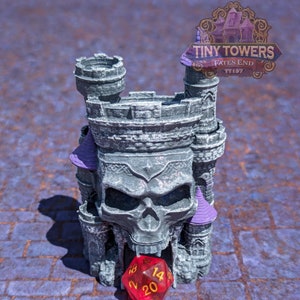 Könnte beinhalten: Ein graues Miniatur-Schloss mit Totenkopf-Design, lila Akzenten und dem Text "Tiny Towers Fates End TT197". Ein roter zwanzigseitiger Würfel liegt vor dem Schloss. Das Schloss steht auf einer lila-braunen Oberfläche.