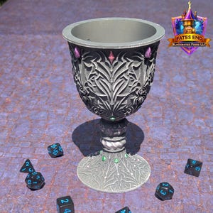 Könnte beinhalten: Ein silberner Kelch mit aufwendigen floralen Mustern und Edelsteinakzenten. Mehrere schwarze Würfel mit blauen Zahlen liegen darum herum. Der Text "Fates End" ist ebenfalls enthalten.