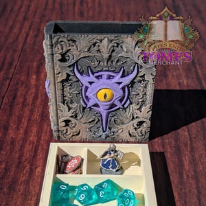 Puede incluir: Una caja de madera blanca con un compartimento para dados y una figura en miniatura. La caja está decorada con un diseño ornamentado marrón oscuro y un símbolo de ojo morado. El texto "Fates End 2024-2025 Tomes Merchant" está impreso en la caja.