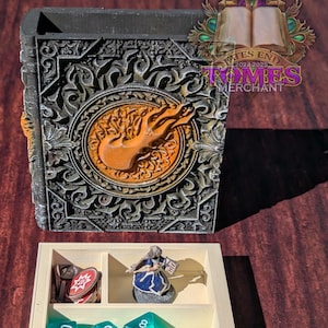 Puede incluir: Una caja de madera blanca con un compartimento para dados, fichas y una figura en miniatura. La caja tiene una tapa decorativa con un diseño negro y naranja y el texto "Fates End Tomes Merchant 2024-2025".