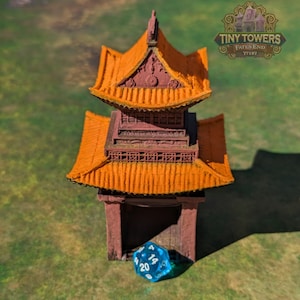 May include: A blue and clear plastic twenty-sided die with white numbers sits in front of a miniature orange and brown pagoda-style building. The building has a sign that reads "Tiny Towers Fates End TT1197".