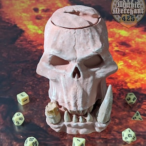 May include: A 3D printed skull-shaped dice tower with a removable lid. The tower is painted in a light pink color and has a detailed skull design. The tower is surrounded by several dice, including a d4, d6, d8, d10, d12, and d20.