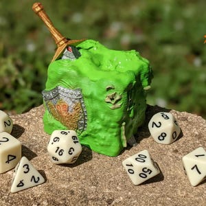 May include: A green, textured dice box with a sword and shield design, surrounded by ivory dice with black numbers. The dice box has a fantasy theme, perfect for tabletop gaming.