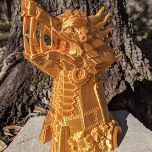 May include: A gold-coloured, 3D printed dragon head dice tower with a detailed, intricate design. The tower is made of plastic and has a textured surface. The tower is designed to hold dice and roll them out of the dragon's mouth.