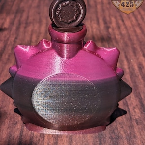 May include: A dark pink and black bottle with a spiky design and a round, dark brown stopper. The bottle has a silver-colored circle on the front. The bottle is sitting on a wooden surface.