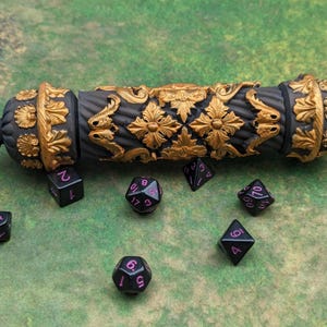 May include: A black and gold decorative dice roller with ornate floral designs. Several black dice with pink numbers are scattered around the roller. The dice roller is on a green surface.