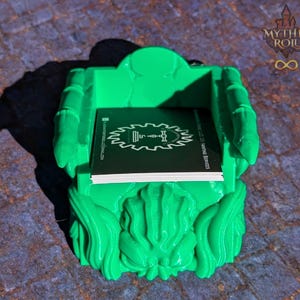May include: A bright green 3D printed throne-shaped business card holder. The holder has a detailed design with a raised, ornate base and a small, rectangular space for holding business cards. The business card holder is sitting on a textured, green surface.