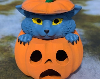 Puede incluir: Una figurita de gato azul con ojos amarillos asoma de una calabaza naranja brillante. La calabaza tiene una cara de calabaza tallada.