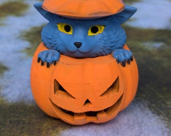 Puede incluir: Una figurita de gato azul asomándose de una calabaza naranja tallada. La calabaza tiene una cara de jack-o'-lantern.