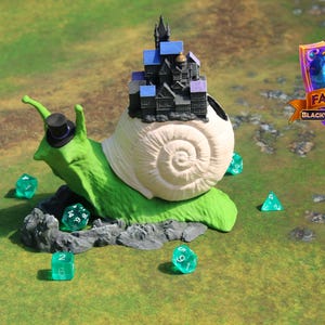 May include: A whimsical green snail figurine wearing a top hat and carrying a miniature castle on its back. Several translucent green dice are scattered around the snail on a green surface. The image includes a logo that says "Fates End".