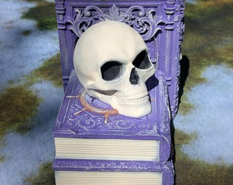 Puede incluir: Un libro morado con una calavera en la parte superior. El libro tiene un diseño detallado y está apilado sobre otro libro.