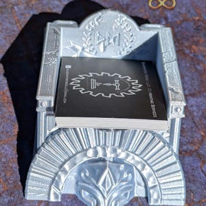 May include: A silver 3D printed business card holder with a decorative design. The holder features a helmet with a crest and a laurel wreath. The holder is sitting on a textured surface.