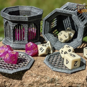 May include: Two gray, octagonal, 3D printed dice towers with a cage design. One tower is open and has white dice inside. The other tower is closed and has purple dice inside. There are several white and purple dice on a gray, octagonal, 3D printed tray. The text "FATES END DICE TOWERS" is on the side of one of the towers.