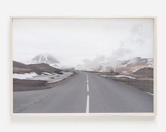 May include: A long, straight, paved road stretches into the distance, with a snow-capped mountain in the background. The road is flanked by a barren, rocky landscape with steam rising from the ground.