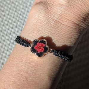 Könnte beinhalten: Ein schwarzes geflochtenes Armband mit einem goldfarbenen Blumenanhänger, in dem eine rote gepresste Blume zu sehen ist.