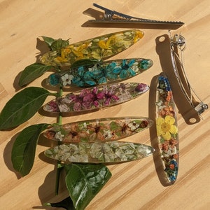 May include: Assortment of seven hair clips featuring pressed flowers encased in clear resin. The clips are oval shaped and come in a variety of colours, including yellow, blue, pink, and white.