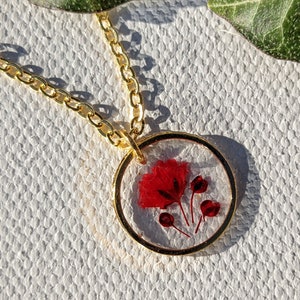 Puede incluir: Un collar de cadena dorada con un colgante redondo que presenta una flor roja prensada dentro de resina transparente.