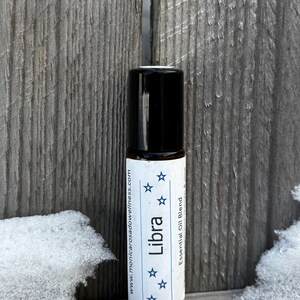 May include: A small, black roll-on bottle with a white label that says "Libra" and "Essential Oil Blend". The label also includes five blue stars and the website address "www.monicarosadowellness.com". The bottle is sitting on a wooden surface with snow in the background.