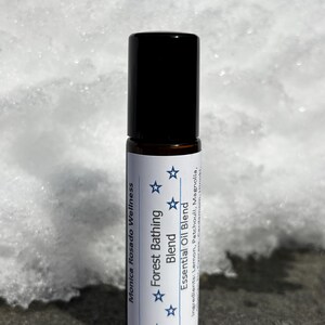 May include: A small, clear glass bottle with a black roll-on top. The label is white with black text that reads "Monica Rosado Wellness Forest Bathing Blend Essential Oil Blend" and has five blue stars. The ingredients are listed as "Lemon, Patchouli, Magnolia, Hinaki".