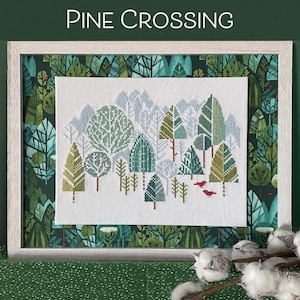 Op de afbeelding: Een kruissteekpatroon met de titel "Pine Crossing" met een winterse bos scène met groene, blauwe en witte bomen. Het patroon is ingelijst in een wit frame met een groene en blauwe gepatroneerde rand. De afbeelding is omgeven door groene stof met witte wattenbolletjes.