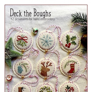 Pode incluir: Doze ornamentos de feltro costurados à mão com vários temas natalinos, incluindo um floco de neve, uma rena, uma meia, um bastão de caramelo, um sino, uma luva, uma coroa, uma árvore, uma bota e um ramo de azevinho. Os ornamentos são enfiados em um barbante vermelho e branco e têm sinos dourados. O título "Deck the Boughs" e o texto "12 ornaments for hand embroidery" estão impressos na parte superior da imagem. O texto "by Kathy Schmitz" está impresso na parte inferior da imagem.