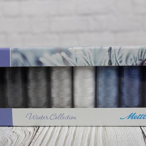 May include: A box set of Mettler Winter Collection thread spools. The spools are in shades of gray and blue. The box has a winter-themed design and the words "Winter Collection" are printed on the front.