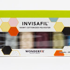 Può includere: Una scatola di fili speciali INVISAFIL. La scatola è bianca con un motivo geometrico di esagoni gialli, neri, rosa, arancioni e blu. Sei rocchetti di filo sono visibili in tonalità di grigio, beige, bordeaux, verde oliva e blu.