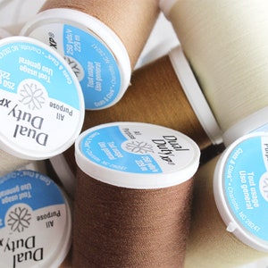 May include: Close-up of several spools of Dual Duty XP thread. The spools are white, brown, and beige. The spools are labeled with the text "Dual Duty XP" and "All Purpose". The spools are 250 yards/229 meters.
