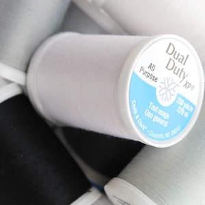 May include: Close-up of a white spool of Dual Duty xp All Purpose thread with a blue label. The label reads "250 yds/y 229 m", "Tout usage Uso general", "Coats & Clark", and "Charlotte, NC 28247".