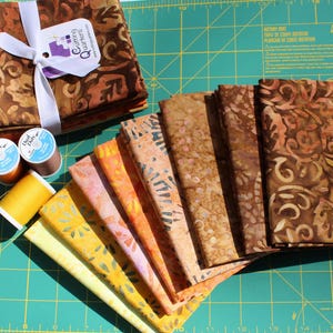 May include: A collection of fabric squares in various warm colors, including brown, orange, and yellow, with floral and abstract patterns. The fabric is bundled with a white ribbon and a tag that reads "Cutting Quarters". Also included are spools of thread.