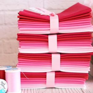 Può includere: Pila di quadrati di tessuto rosa, rosso e bianco legati con un nastro rosa. Il tessuto è piegato e impilato in un motivo sfumato. Ci sono due bobine di filo in primo piano, una rosa e una rossa.