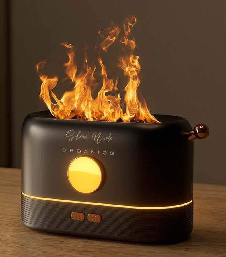 May include: A matte black, rectangular diffuser with a rounded top emitting a realistic flame effect. The front features the gold text "Shere' Nicole ORGANICS", a circular yellow light, and two small buttons. A copper-colored lever is on the side.