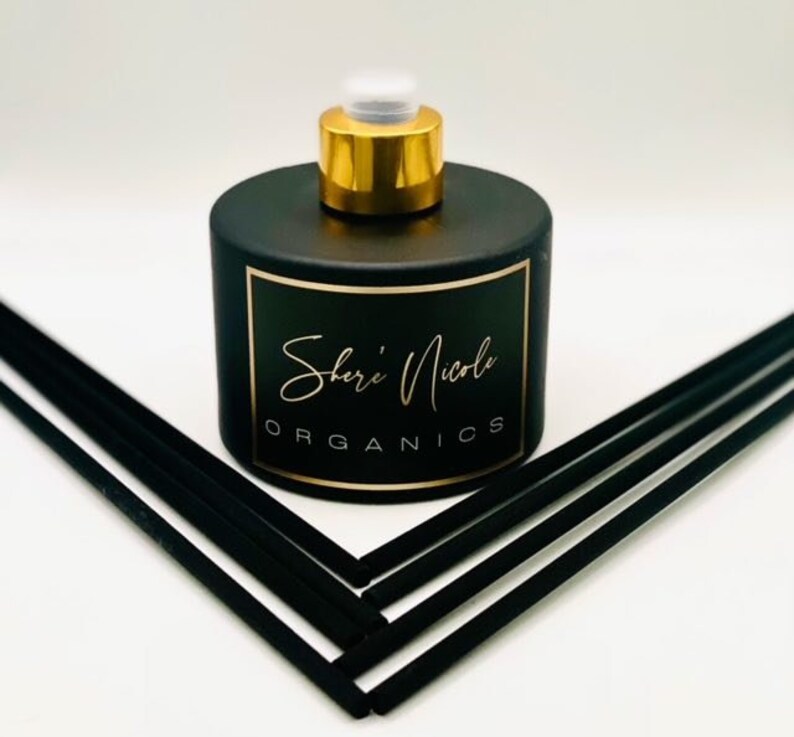 May include: A matte black cylindrical reed diffuser bottle with a gold-colored cap. The label features the text Shere Nicole ORGANICS in gold lettering. Six black reed diffuser sticks are arranged in a V-shape pointing toward the bottle.