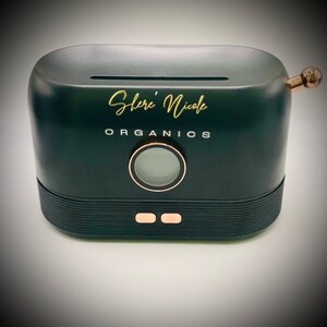 May include: A matte black, oval-shaped electronic device with a top slot, featuring the gold text "Shere' Nicole" and "ORGANICS" printed on the front. It includes a circular display, two rose gold buttons, and a rose gold knob on the right side.